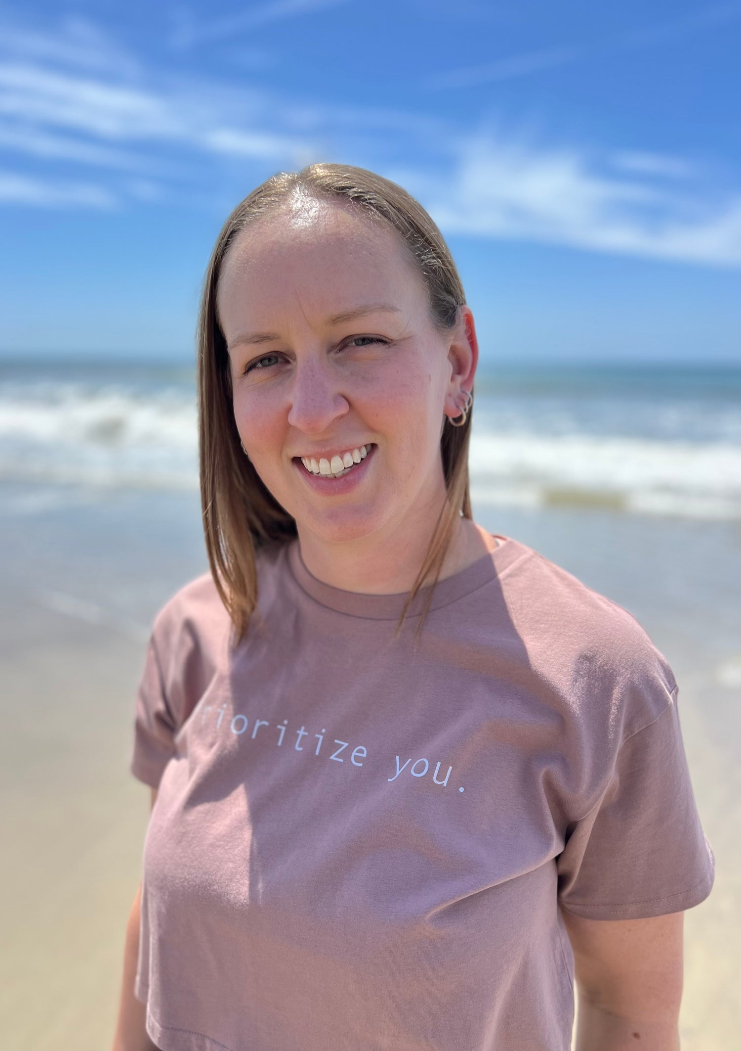 Girl smiling in a cropped t-shirt that says "Prioritize you" in front of a beach